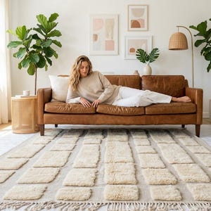 Peut inclure: Un salon avec un canapé en cuir marron, un tapis beige à motifs rectangulaires et des œuvres d'art abstraites encadrées. Un lampadaire avec un abat-jour tissé et des plantes en pot complètent le décor. Le tapis a une bordure frangée.