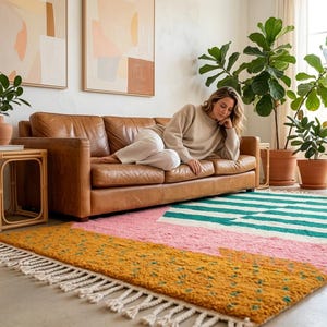 Peut inclure: Un tapis rectangulaire au design color-block composé de sections jaune moutarde, rose pâle et blanc avec des rayures bleu sarcelle. Le tapis présente un poil texturé et touffu, agrémenté de franges blanches épaisses et nouées sur les bords.