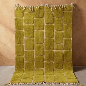 Peut inclure: Un tapis rectangulaire vert avec un motif texturé en forme de blocs. Le tapis présente un fond beige clair avec des sections carrées en relief d'un vert vif. Les bords sont finis avec une frange de pompons blancs, ajoutant une touche décorative.