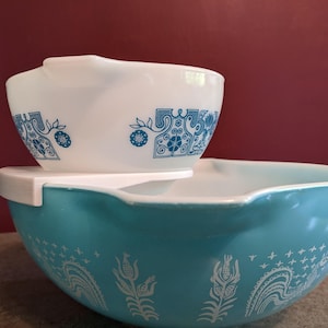 May include: Two vintage Pyrex mixing bowls. The larger bowl is turquoise with white floral and paisley designs. The smaller bowl is white with blue floral patterns. Both bowls have a classic, retro aesthetic.