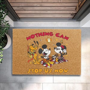 May include: A rectangular doormat with a cartoon image of Mickey and Minnie Mouse, Pluto, and a bird. The text "NOTHING CAN STOP US NOW" is in red and yellow. The mat is brown.