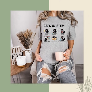 May include: A grey t-shirt with the words "CATS IN STEM" and images of black cats illustrating scientific concepts. The top has a crew neck and short sleeves.