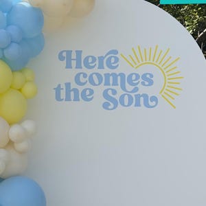 May include: A white arch backdrop with the text "Here comes the Son" in yellow and blue lettering. The backdrop is decorated with a garland of yellow, white, and blue balloons.