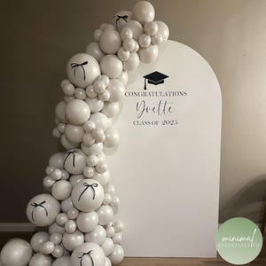 May include: A white arched backdrop featuring a black graduation cap icon and the text CONGRATULATIONS Yvette CLASS OF 2025. A large cluster of white balloons is attached to the left side, with several balloons accented by small black ribbons.