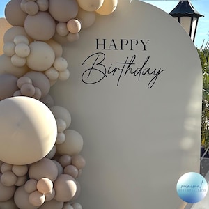 May include: A white backdrop with the text "HAPPY Birthday" in black script. The backdrop is decorated with a garland of beige and cream balloons.