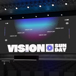 Peut inclure: Une scène avec un grand écran affichant "VISION DAY" en blanc, avec un dégradé bleu et violet. La scène est installée dans un auditorium avec des rangées de sièges. Le texte "VISION DAY" est répété.
