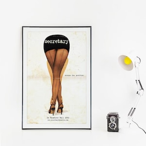 Può includere: Un poster incorniciato con la parola "secretary" sopra una raffigurazione delle gambe di una persona con calze nere e tacchi alti. Il poster include il testo "Assume the position" e "In Theatres Fall 2002". Una fotocamera vintage e una lampada da scrivania bianca sono sulla superficie bianca.