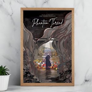 Può includere: Una stampa artistica incorniciata con il titolo del film "Phantom Thread" in una scrittura elegante. L'opera d'arte raffigura una coppia che si abbraccia in un'ambientazione colorata e onirica, con una cornice in legno marrone chiaro. La stampa è esposta su una superficie bianca.
