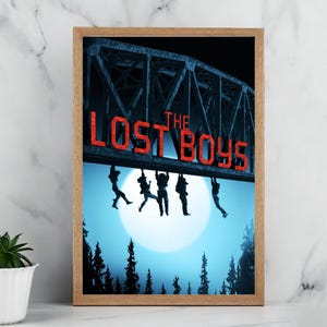 May include: Framed print with the text "THE LOST BOYS" in red above silhouetted figures hanging from a bridge. The background depicts a full moon and dark trees, creating a dramatic scene.