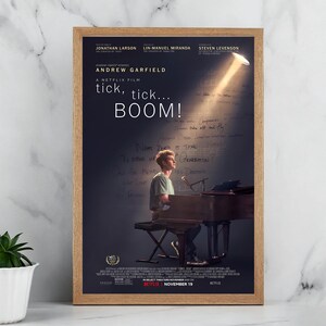 Może przedstawiać: Oprawiony plakat filmowy do filmu "tick, tick... BOOM!" przedstawia Andrew Garfield przy fortepianie. Plakat ma ciemne tło z odręcznymi notatkami i reflektorem. Tytuł filmu jest dużymi białymi literami.