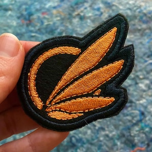 May include: Embroidered patch with a black background and a gold-colored design. The design features a stylized wing or leaf shape with a circular element. The patch has a black border and is held by a hand.