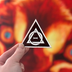 May include: A triangular embroidered patch with a black background and white detailing. The design includes a white border, a circle with a line, and the letter "I". The patch is held up.