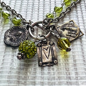 May include: A silver-toned necklace with multiple charms. The necklace features a double chain with green beads. Charms include religious medallions, a heart-shaped pendant, and a cluster of green crystal beads. The background is a white, textured surface.