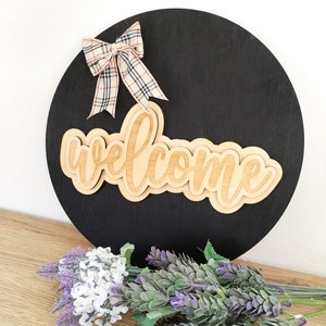 Puede incluir: Un letrero circular de madera negra con la palabra welcome en letras de madera clara. Un lazo de cinta a cuadros con líneas beige, negras y rojas está sujeto en la parte superior izquierda. El letrero se muestra con ramas de lavanda morada y blanca.