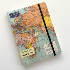 May include: A rectangular notebook featuring a vintage world map print in shades of orange, yellow, and light blue. The cover includes a black elastic closure, a blue 'BY AIR MAIL PAR AVION' label, and a yellow 'VINTAGE MAP NOTEBOOK' sticker.