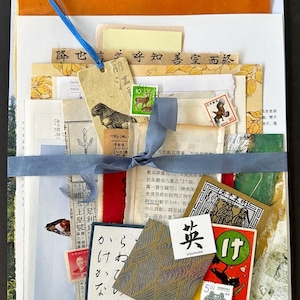 May include: A stack of vintage papers and ephemera tied with a blue ribbon. The papers are in various shades of brown, yellow, and green, and feature Japanese characters, stamps, and illustrations. The papers are tied together with a blue ribbon and a tag that says "Courage".