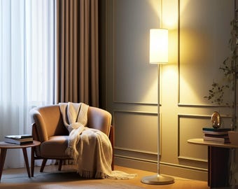 May include: A modern floor lamp with a cylindrical white shade illuminates a cozy living space. A tan armchair with a cream-colored throw blanket sits near a small wooden side table. The room features neutral tones and a warm, inviting atmosphere.