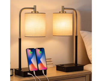 May include: Two matching bedside lamps with a black base and gold accents. Each lamp has a beige fabric shade and a curved arm. Two smartphones are charging on the base of one lamp.