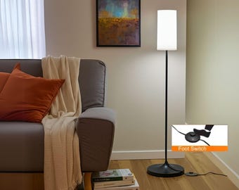 May include: A black floor lamp with a white rectangular lampshade. The lamp has a round base and a foot switch. The lamp is next to a gray sofa with orange pillows and a cream-colored throw blanket.