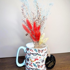 May include: A white ceramic mug with a light blue handle and rim, decorated with colorful butterfly illustrations. The mug holds a bouquet of dried flowers in red, white, and yellow. A black tag with the words "made with LOVE" hangs from the mug.
