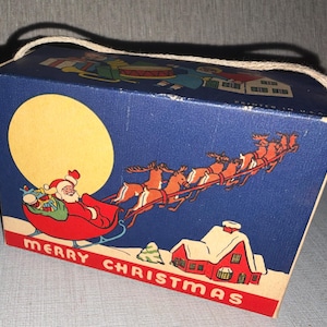 May include: A vintage Christmas gift box with a blue background featuring Santa Claus in a sleigh pulled by reindeer, a large yellow moon, and a red house. The box has a rope handle and the words "MERRY CHRISTMAS" in red.