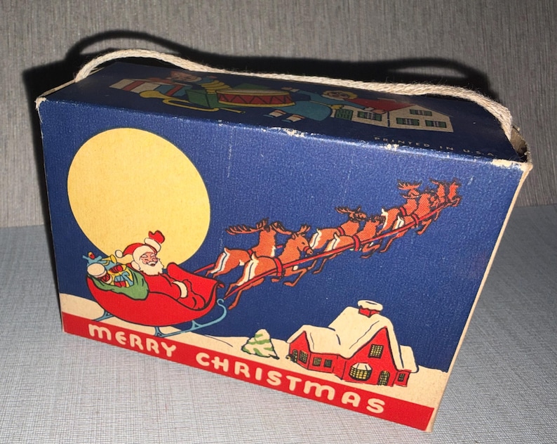 May include: A vintage Christmas gift box with a blue background featuring Santa Claus in a sleigh pulled by reindeer, a large yellow moon, and a red house. The box has a rope handle and the words "MERRY CHRISTMAS" in red.