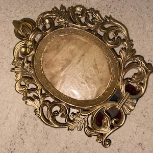 May include: An oval-shaped gold-toned metal picture frame featuring an intricate, ornate border with scrolling leaf and floral motifs. The center contains a clear protective pane over a textured, light beige backing material with a visible pattern.