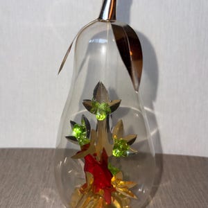 May include: Clear glass pear-shaped ornament with a gold-colored metal top and accents. Inside, a decorative arrangement features gold, red, and green elements, including what appears to be a small tree with green gemstone accents. A clear crystal bead hangs from the top.