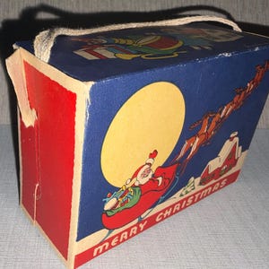 May include: Vintage Christmas-themed box with a red and blue color scheme. The box features a Santa Claus illustration in a sleigh with reindeer, a large yellow moon, and the words "MERRY CHRISTMAS". A rope handle is attached to the top.