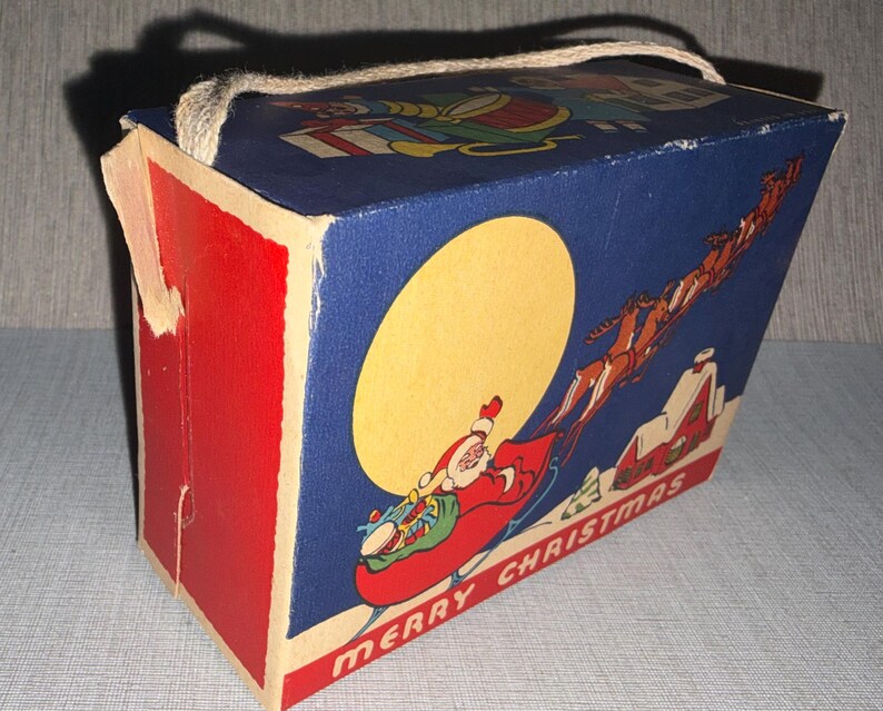 May include: Vintage Christmas-themed box with a red and blue color scheme. The box features a Santa Claus illustration in a sleigh with reindeer, a large yellow moon, and the words "MERRY CHRISTMAS". A rope handle is attached to the top.