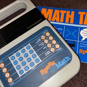 May include: A vintage Speak & Math educational toy by Texas Instruments. The gray and blue device features number buttons and function keys. A blue and orange Math Talk activity book is also visible, with the text "sixteen hundred one."