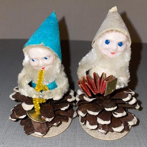 May include: Two vintage Christmas elf figurines made of white cotton and sitting on pine cones. One elf is wearing a blue hat and playing a yellow saxophone. The other elf is wearing a white hat and playing a red accordion.