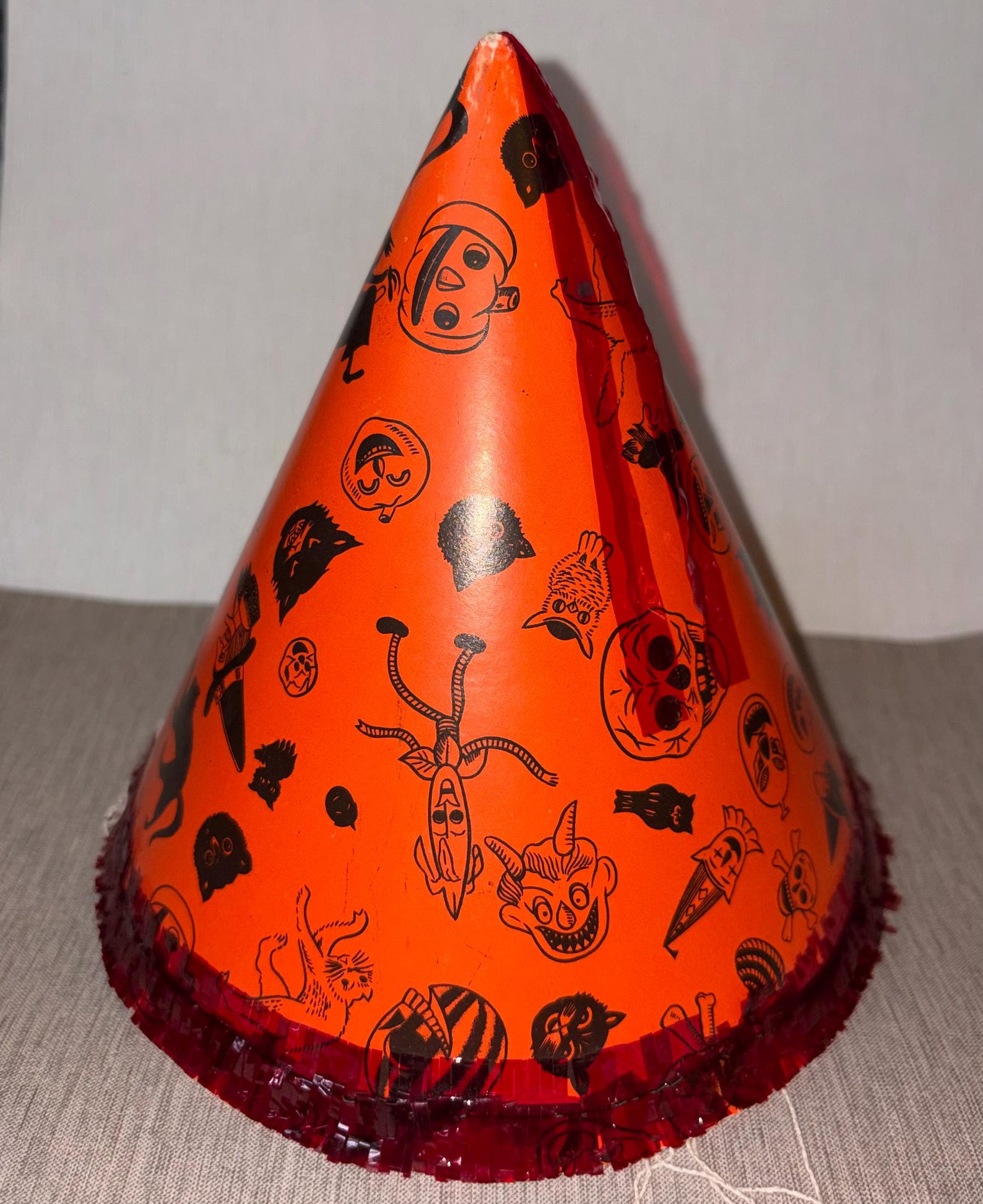 A Patterned Cone with a Dark Fringe Base Vintage orange cone hat with small black Halloween characters and dark fringe around the rim.