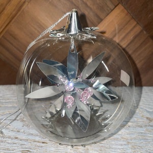 May include: Clear glass Christmas ornament with a silver star-shaped design and pink beads inside.
