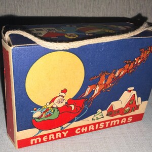 May include: Vintage Christmas-themed box with a blue background featuring Santa Claus in a sleigh, reindeer, a full moon, and a snow-covered house. The box has red sides and the words "MERRY CHRISTMAS" at the bottom.