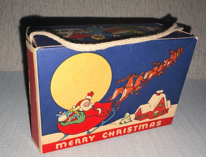 May include: Vintage Christmas-themed box with a blue background featuring Santa Claus in a sleigh, reindeer, a full moon, and a snow-covered house. The box has red sides and the words "MERRY CHRISTMAS" at the bottom.