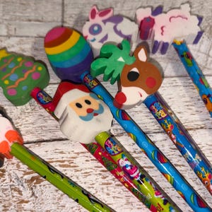 May include: A collection of colorful pencils with decorative erasers. The erasers feature various designs, including a penguin, Santa Claus, a Christmas tree, a rainbow egg, a bunny, a reindeer, and a sheep.