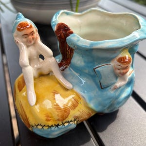 May include: A ceramic planter shaped like a shoe with a blue upper and yellow toe. Two small elf-like figures with brown hair are attached to the exterior, one sitting on the toe and one peeking from the side. The interior is glazed in white.