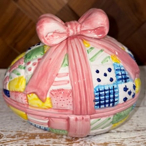 May include: A ceramic Easter egg-shaped trinket box with a pink bow on top. The box is decorated with a patchwork design in pastel colors, including pink, blue, yellow, and green. The lid is secured with a pink ribbon.