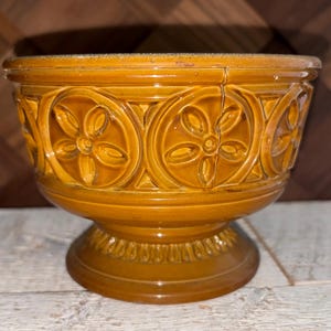 May include: A pedestal bowl in a warm, golden-brown hue. The ceramic bowl features a detailed floral and geometric pattern around the body and base. The rim has a slightly lighter tone, and the bowl sits on a decorative pedestal.