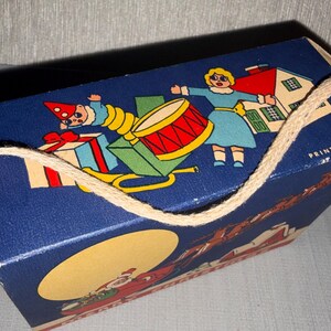 May include: A vintage blue gift box with a rope handle. The box features a colorful illustration of a drum, a house, and figures in festive attire. The text "PRINTED IN U.S.A." is visible.