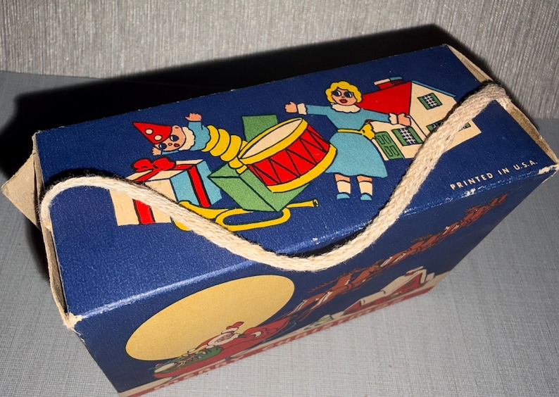May include: A vintage blue gift box with a rope handle. The box features a colorful illustration of a drum, a house, and figures in festive attire. The text "PRINTED IN U.S.A." is visible.