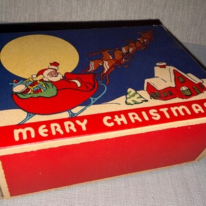 May include: Vintage Christmas box with a festive illustration. Santa Claus in his sleigh, pulled by reindeer, flies across a night sky with a full moon. A red house with snow-covered roof and the words "MERRY CHRISTMAS" are also featured.