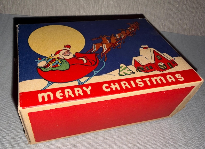 May include: Vintage Christmas box with a festive illustration. Santa Claus in his sleigh, pulled by reindeer, flies across a night sky with a full moon. A red house with snow-covered roof and the words "MERRY CHRISTMAS" are also featured.