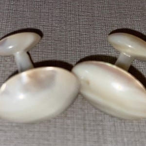 May include: A pair of oval-shaped, pearlescent white cufflinks. Each cufflink has a small, round, dark-colored top connected to an oval base by a short, white stem. The cufflinks are displayed on a textured, gray surface.