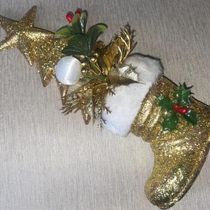 May include: A gold glitter Christmas stocking ornament with a white faux fur cuff. The stocking is decorated with a star, a white ball, and sprigs of holly with red berries. The ornament is designed for holiday decor.