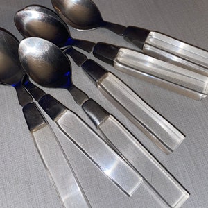 May include: Set of six spoons with stainless steel bowls and clear, faceted acrylic handles. The spoons are arranged on a gray surface, showcasing their reflective qualities and modern design. Ideal for serving or dining.