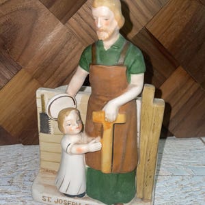 May include: A ceramic figurine of St. Joseph the Workman with a child. St. Joseph is depicted in a green shirt and brown apron, holding a cross. The child is in a white robe. The base reads "ST. JOSEPH THE WORKMAN."