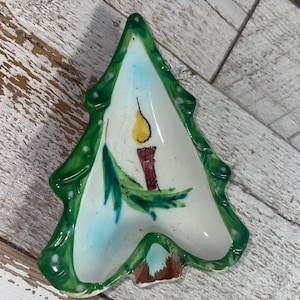 May include: A ceramic Christmas tree-shaped dish with a green border and a white interior. The interior features a candle with a yellow flame, a red base, and green leaves. The dish is approximately 6 inches long.