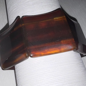 May include: A dark brown, rectangular bracelet with a tortoiseshell pattern. The bracelet is made of a glossy material and has a curved shape. The bracelet is resting on a white, textured surface.